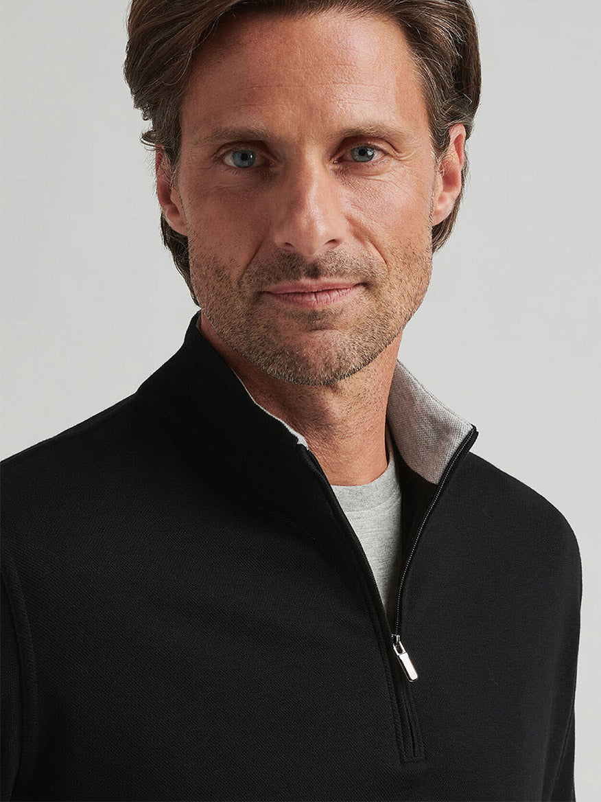 A man with light brown hair and stubble wears the Peter Millar Crown Comfort Pullover in Black over a light grey shirt, standing against a plain light background.