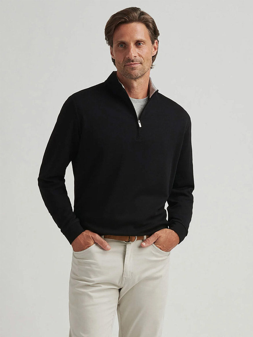A man stands against a plain background, wearing the Peter Millar Crown Comfort Pullover in black over a white shirt, paired with light-colored pants and a tan belt.