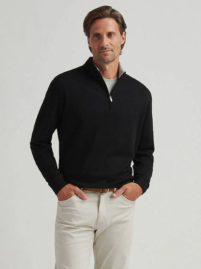 A man stands against a plain background, wearing the Peter Millar Crown Comfort Pullover in black over a white shirt, paired with light-colored pants and a tan belt.