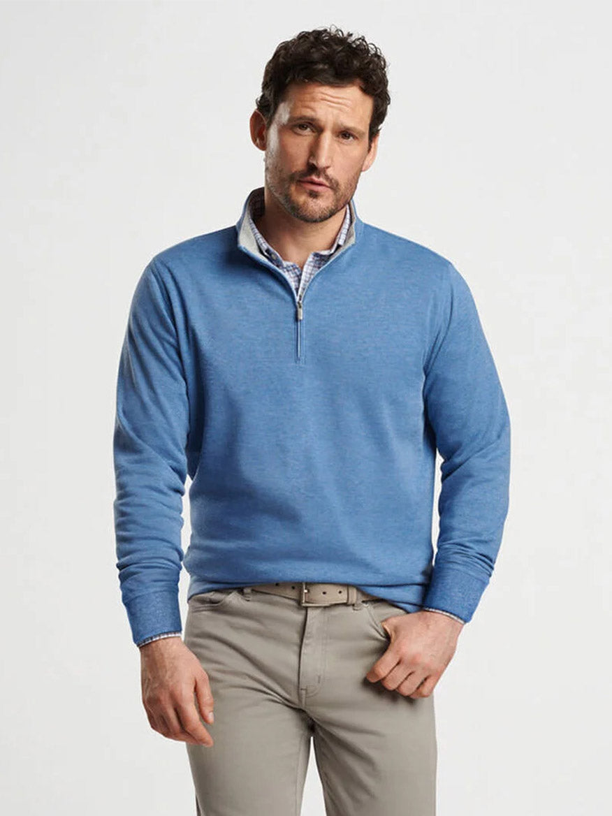 A person with short curly hair and light facial hair is wearing a Peter Millar Crown Comfort Pullover in Astral Blue over a checkered shirt and beige trousers, standing against a plain white background.