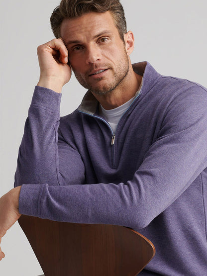 A man with short brown hair and light stubble sits on a wooden chair, resting his head on his hand, wearing the Peter Millar Crown Comfort Pullover in Deep Violet.