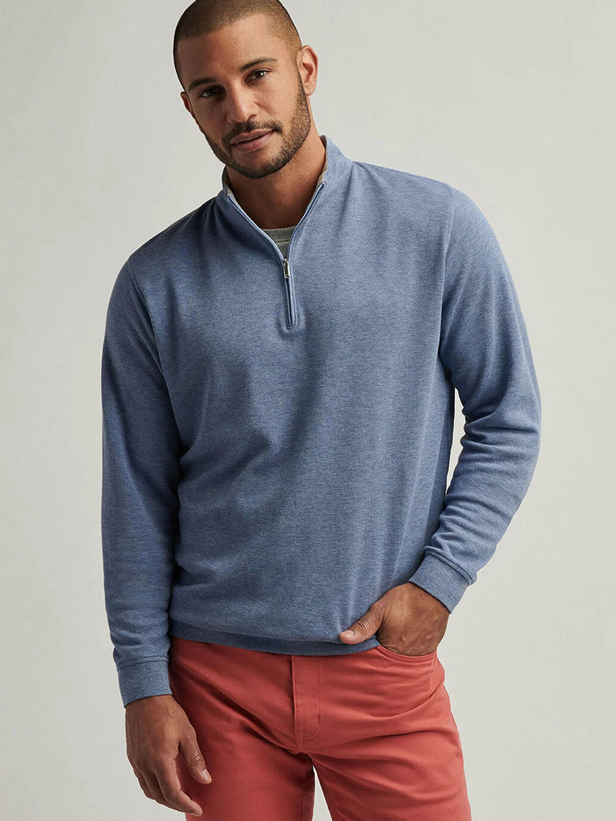 A man models the Peter Millar Crown Comfort Pullover in Blue Freesia, featuring four-way stretch, and pairs it with salmon pants against a light gray backdrop.