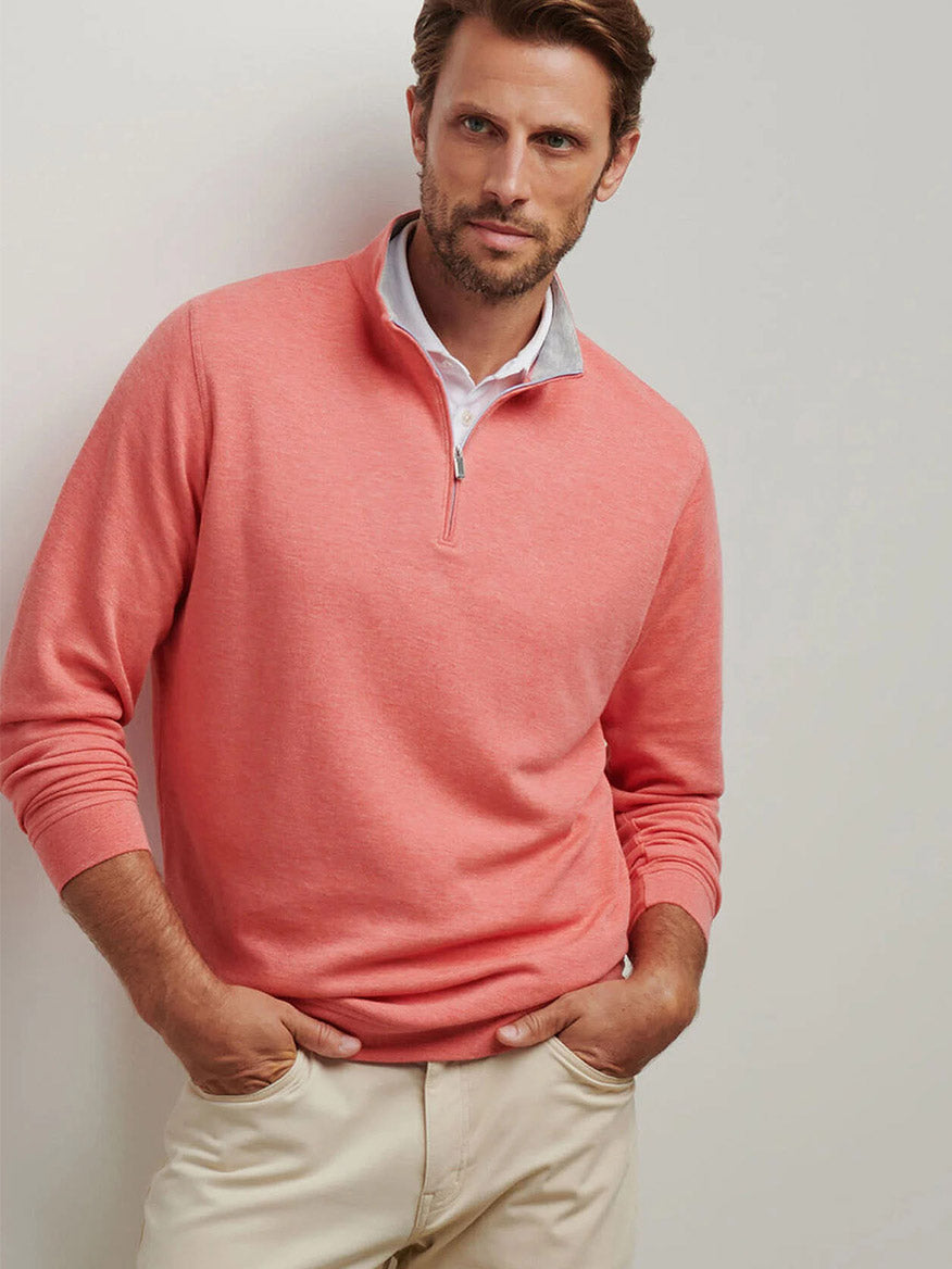 A person wearing the Peter Millar Crown Comfort Pullover in Geranium and beige pants stands with hands in pockets against a neutral background, showcasing its classic fit.