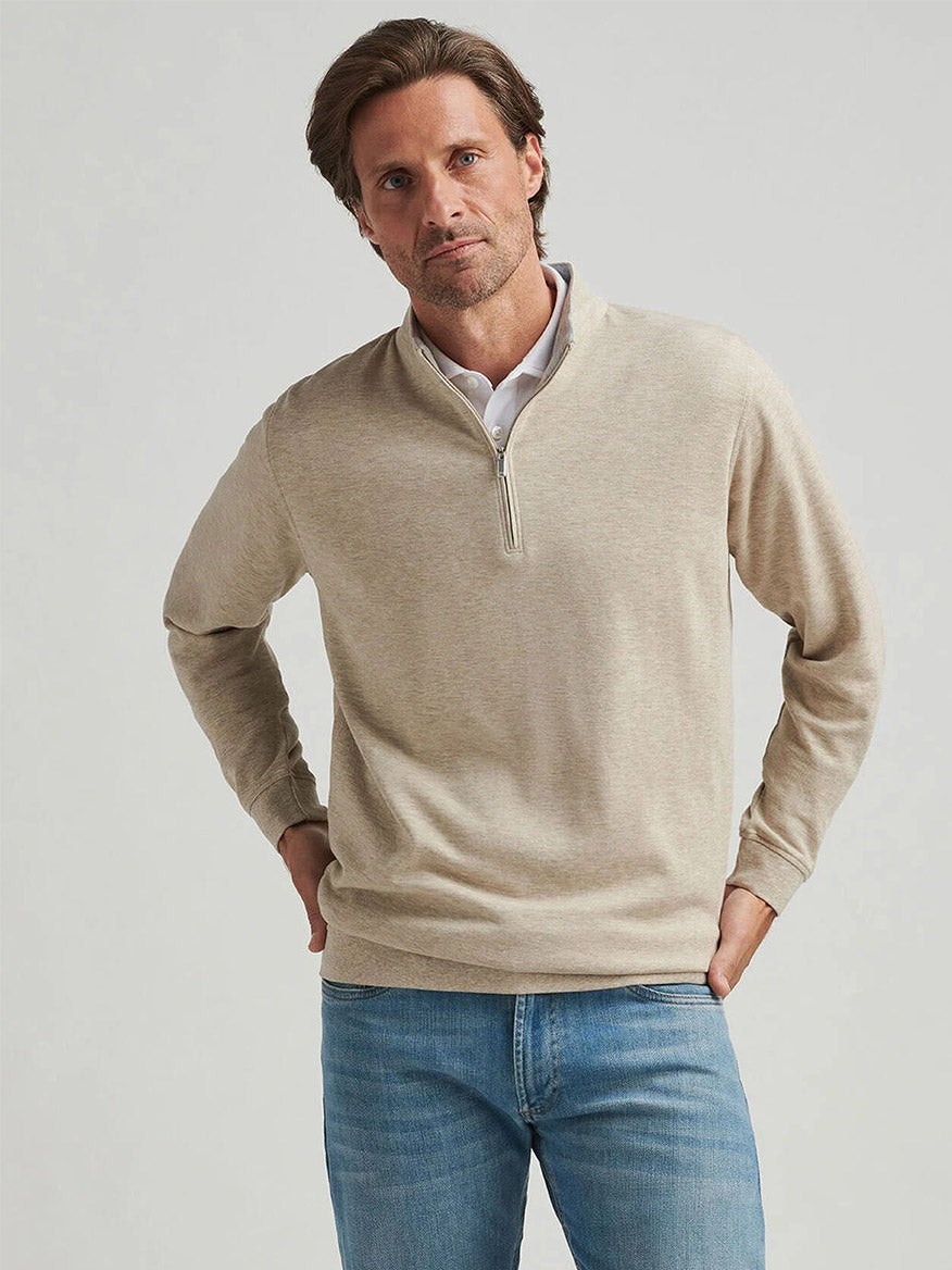 A person in a Peter Millar Crown Comfort Pullover in Stone and blue jeans stands against a plain background.