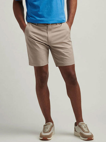 Man wearing a blue classic fit shirt, Peter Millar Crown Comfort Short in Khaki, and beige sneakers stands with hands in pockets against a plain background.