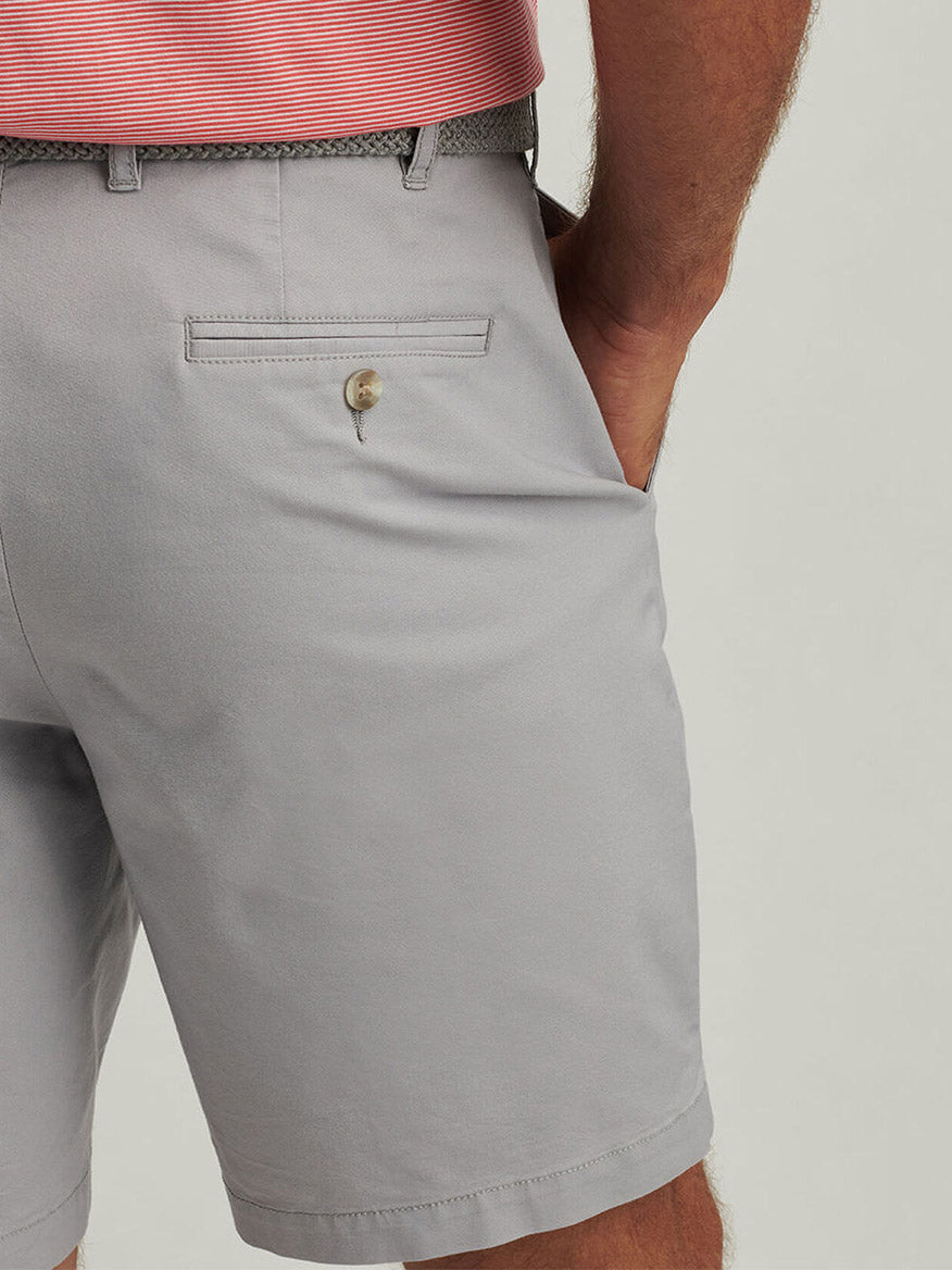 A person wearing Peter Millar Crown Comfort Short in Mountain Grey, featuring four-way stretch, a back pocket with button, a woven belt, and a pink striped shirt, with one hand in the pocket.