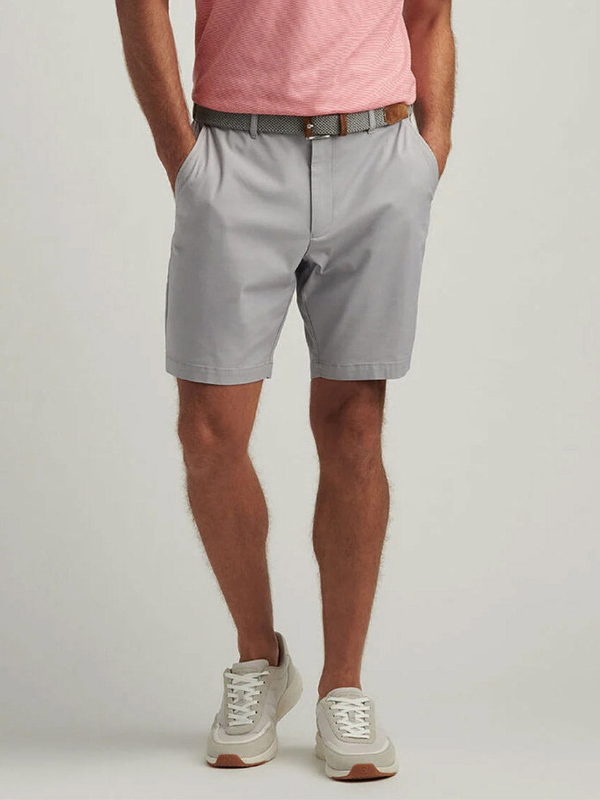 A person stands with hands in pockets against a plain background, wearing Peter Millar Crown Comfort Short in Mountain Grey, a pink shirt, gray belt, and white sneakers.