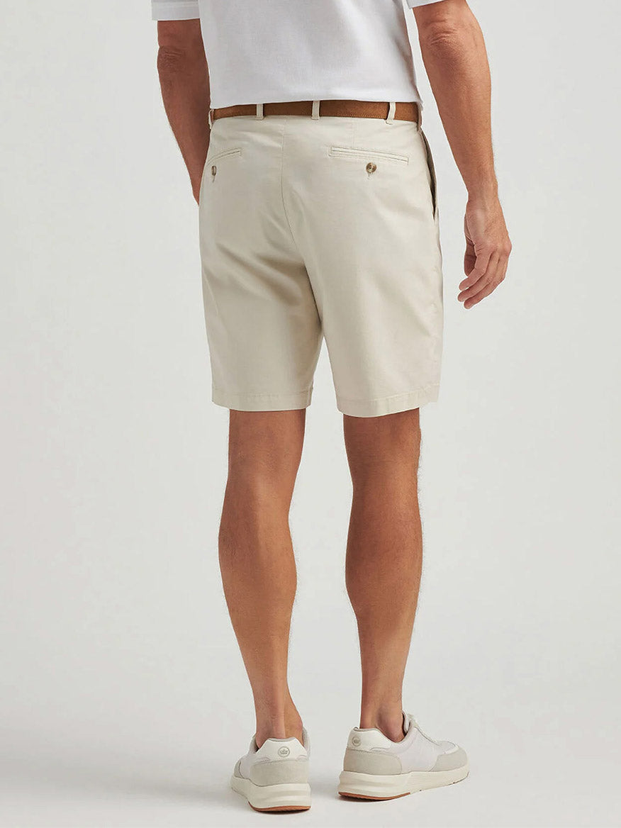 A man stands facing away from the camera, wearing a white polo shirt, the Peter Millar Crown Comfort Short in Stone, a brown belt, and white sneakers.