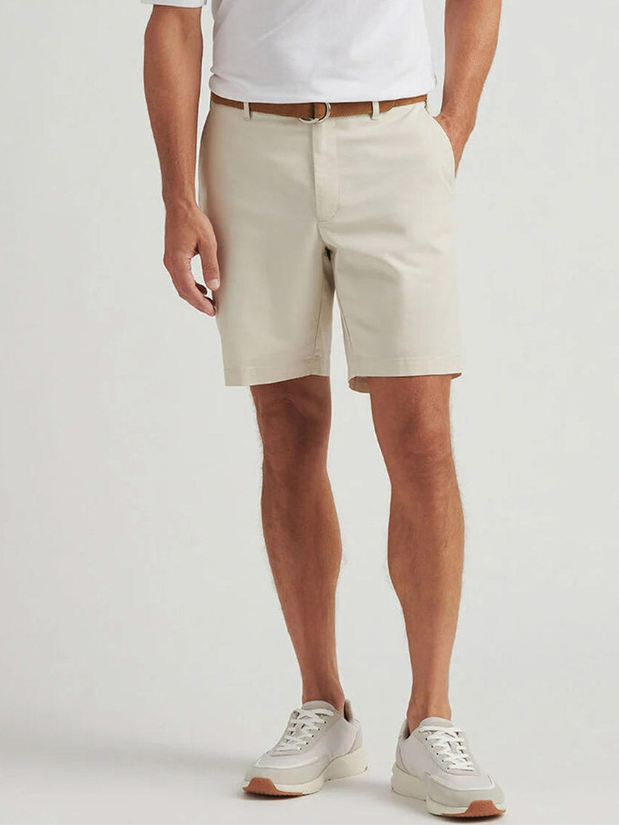 A person wearing a white polo shirt, Peter Millar Crown Comfort Short in Stone made from plain weave fabric for modern mobility, a brown belt, and white sneakers stands against a plain light background.