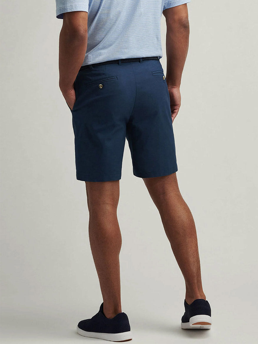 A man stands with his back to the camera, wearing a light blue shirt, black belt, dark blue slip-on shoes, and Peter Millar Crown Comfort Short in Washed Navy crafted from plain weave fabric.