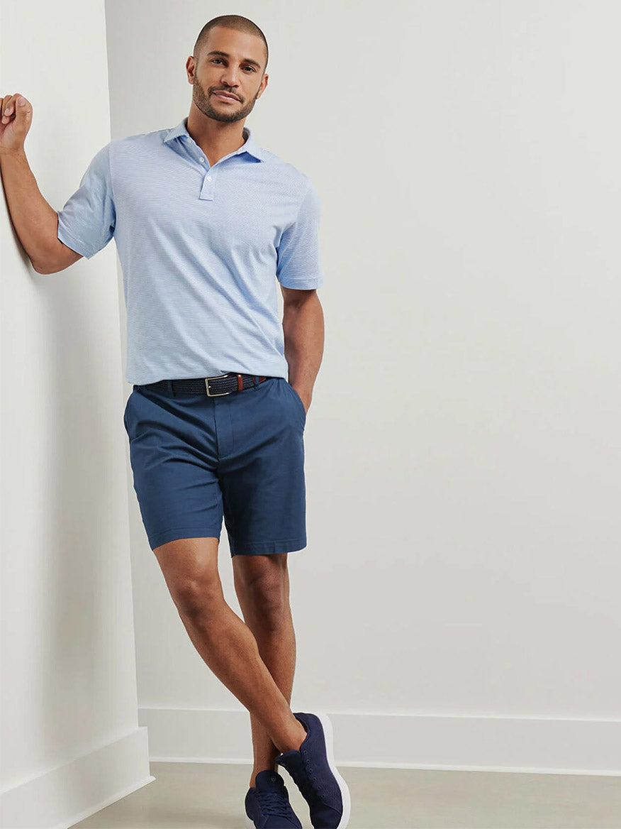 A man stands against a white wall, wearing a light blue polo shirt, a black belt, navy sneakers, and the Peter Millar Crown Comfort Short in Washed Navy made from versatile classics plain weave fabric.