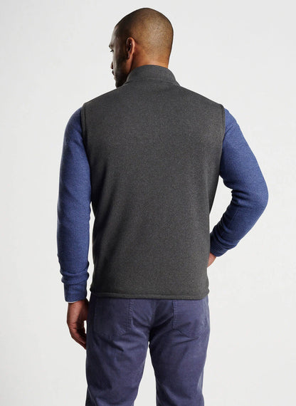 A man is standing with his back facing the camera, wearing an Iron-colored Peter Millar Crown Sweater Fleece Vest over a blue long-sleeve shirt and grey pants, highlighting the use of recycled materials.