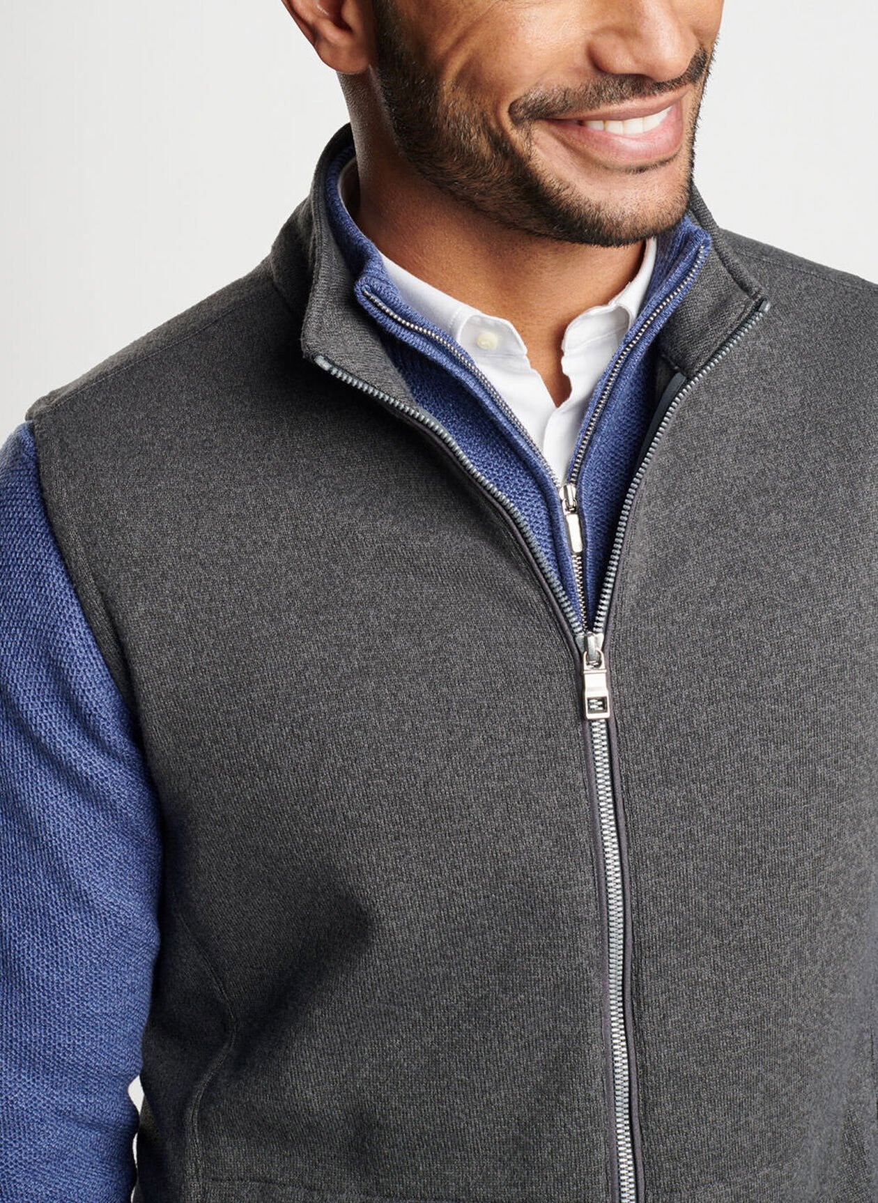 A man is smiling, dressed in a Peter Millar Crown Sweater Fleece Vest in Iron over a blue long-sleeve shirt and a white collared shirt.