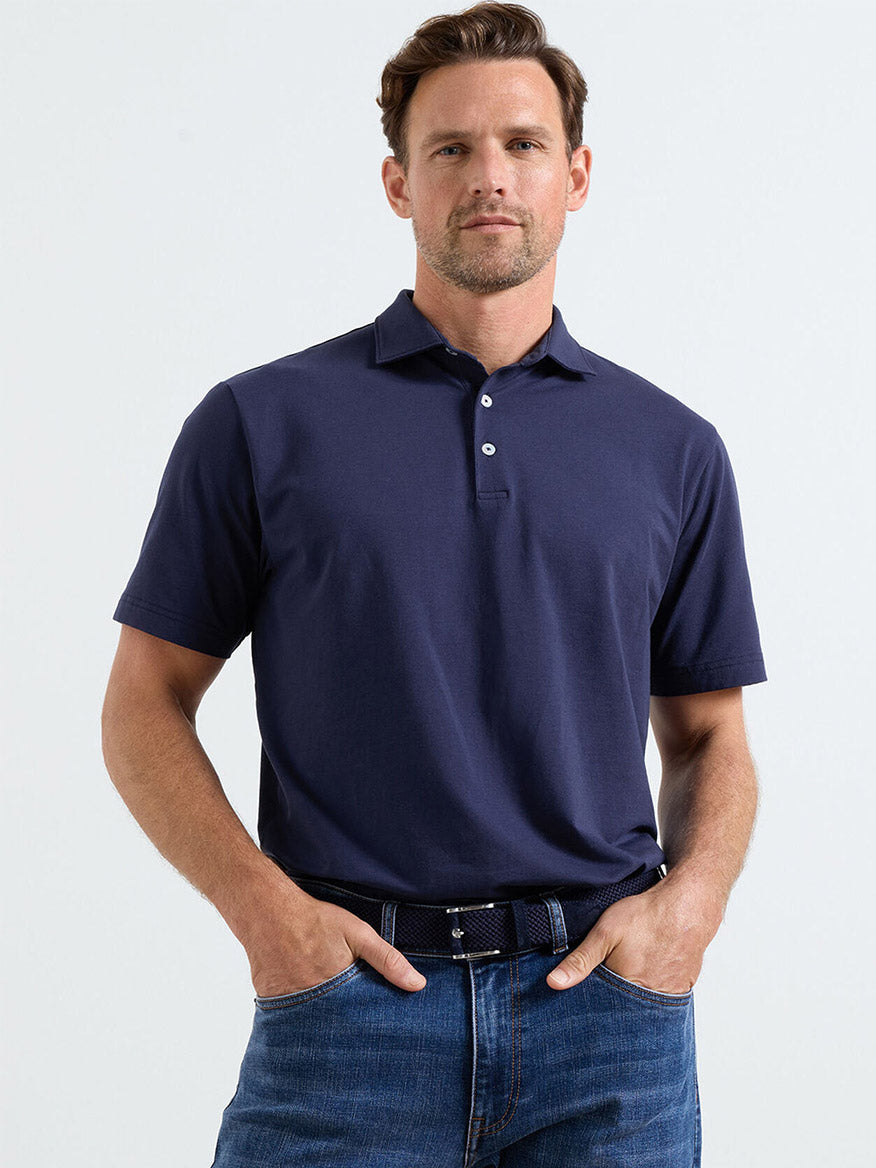Man wearing a Peter Millar Crown Flex Cotton Polo in navy and blue jeans, standing with hands in pockets against a plain light background.