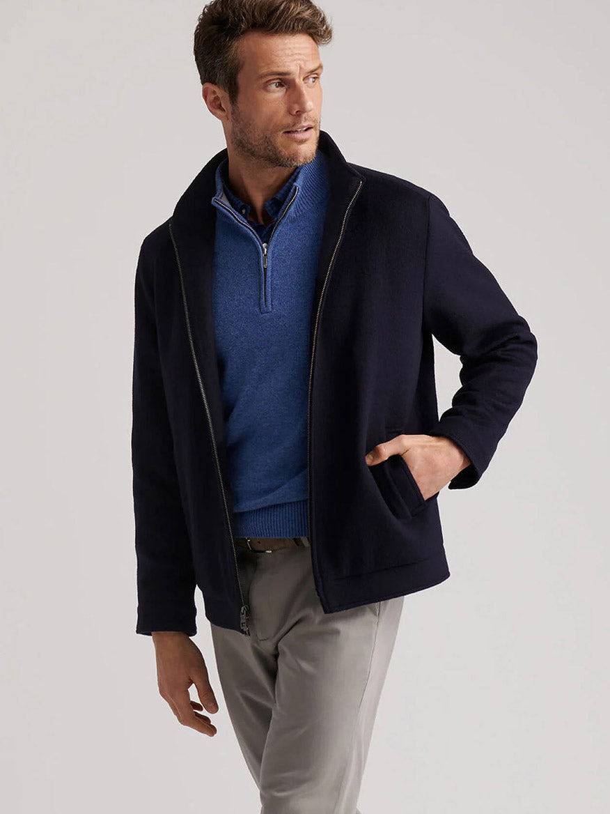 A man wearing the Peter Millar Crown Flex Merino Bomber in Navy stands against a plain background over a blue quarter-zip sweater and light gray pants, looking to the side.