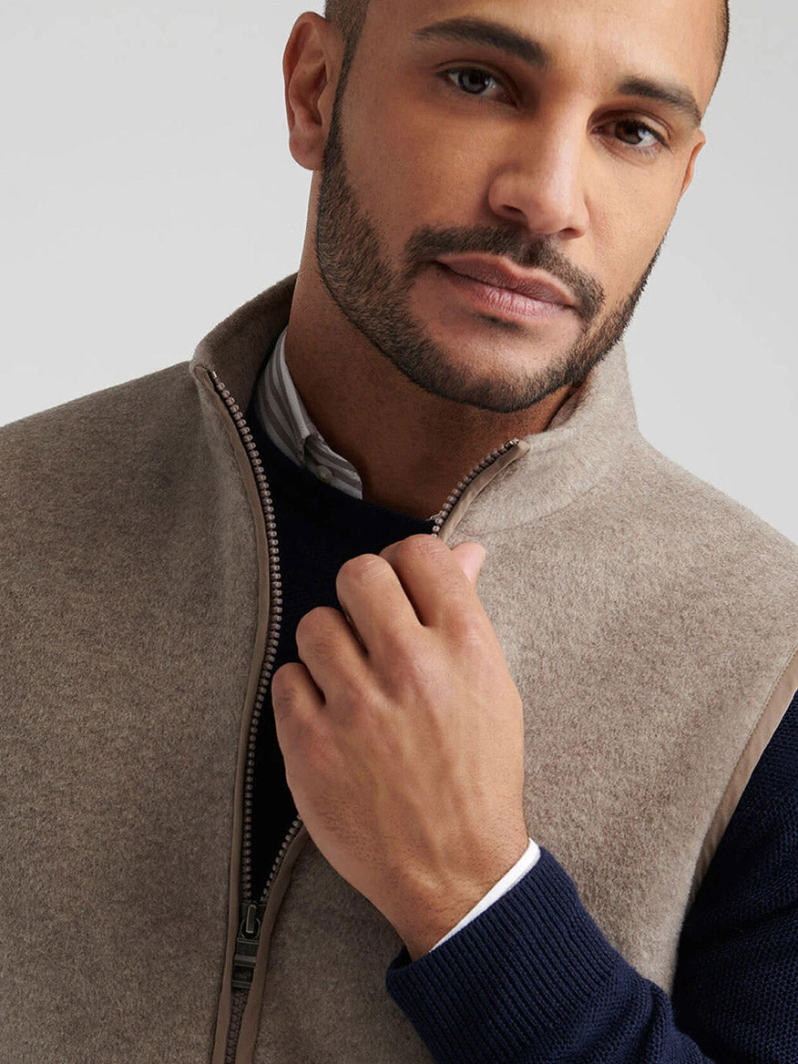 A man wearing the Peter Millar Crown Flex Merino Vest in Grain over a navy sweater and striped shirt looks at the camera while holding the zipper.