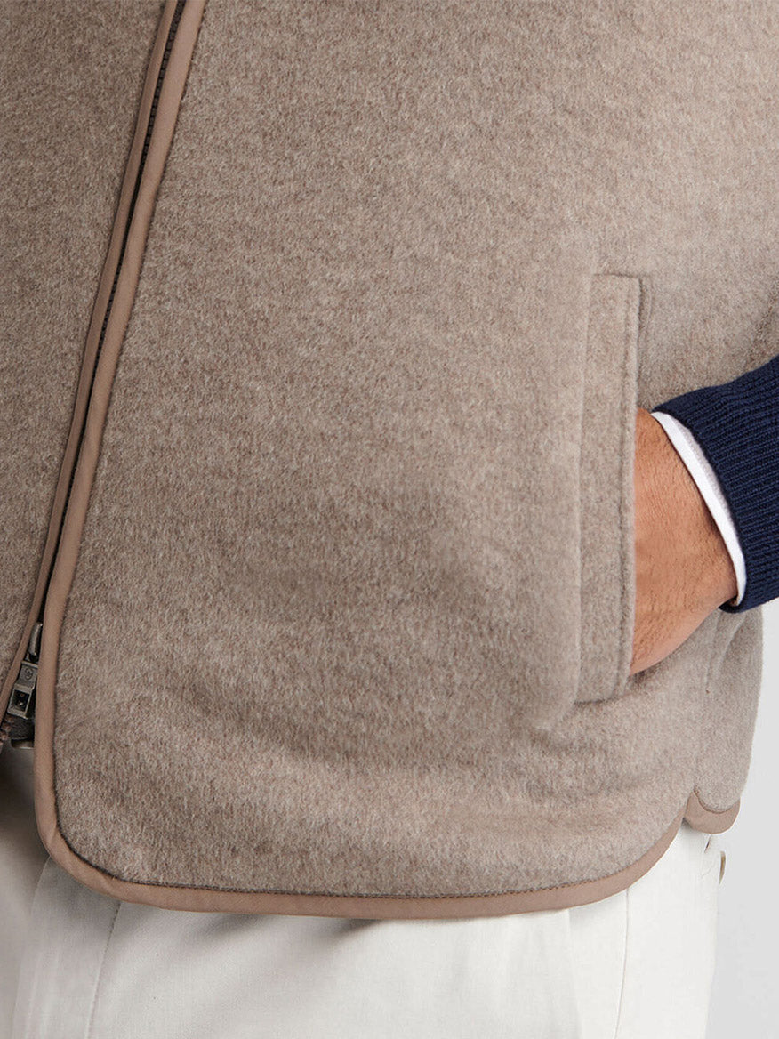 Close-up of a person wearing the Peter Millar Crown Flex Merino Vest in Grain, a beige stretch knit Merino wool vest, layered over a navy sweater and white pants. Their right hand is partially tucked into the vest pocket.