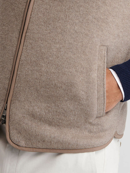 Close-up of a person wearing the Peter Millar Crown Flex Merino Vest in Grain, a beige stretch knit Merino wool vest, layered over a navy sweater and white pants. Their right hand is partially tucked into the vest pocket.