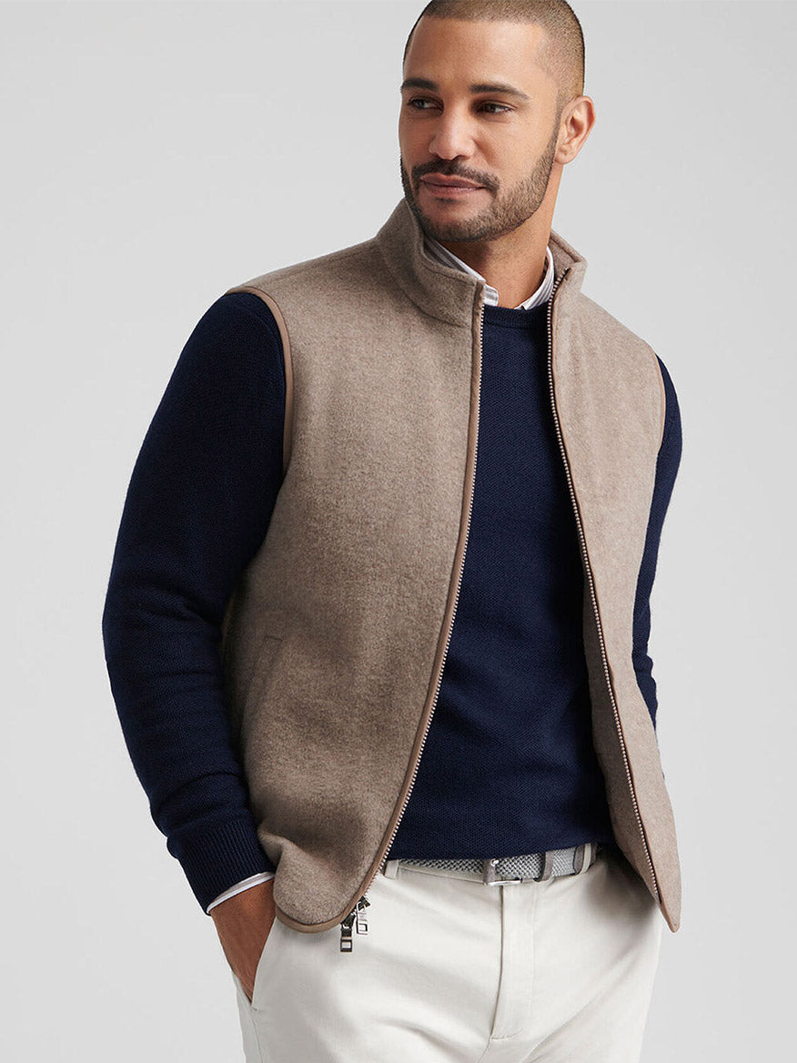 A man wears the Peter Millar Crown Flex Merino Vest in Grain over a navy sweater with white pants, one hand in his pocket—an elegant and versatile layer for any refined look.
