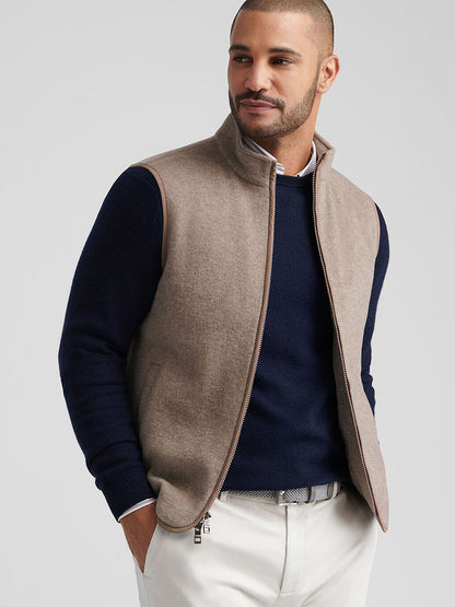 A man wears the Peter Millar Crown Flex Merino Vest in Grain over a navy sweater with white pants, one hand in his pocket—an elegant and versatile layer for any refined look.