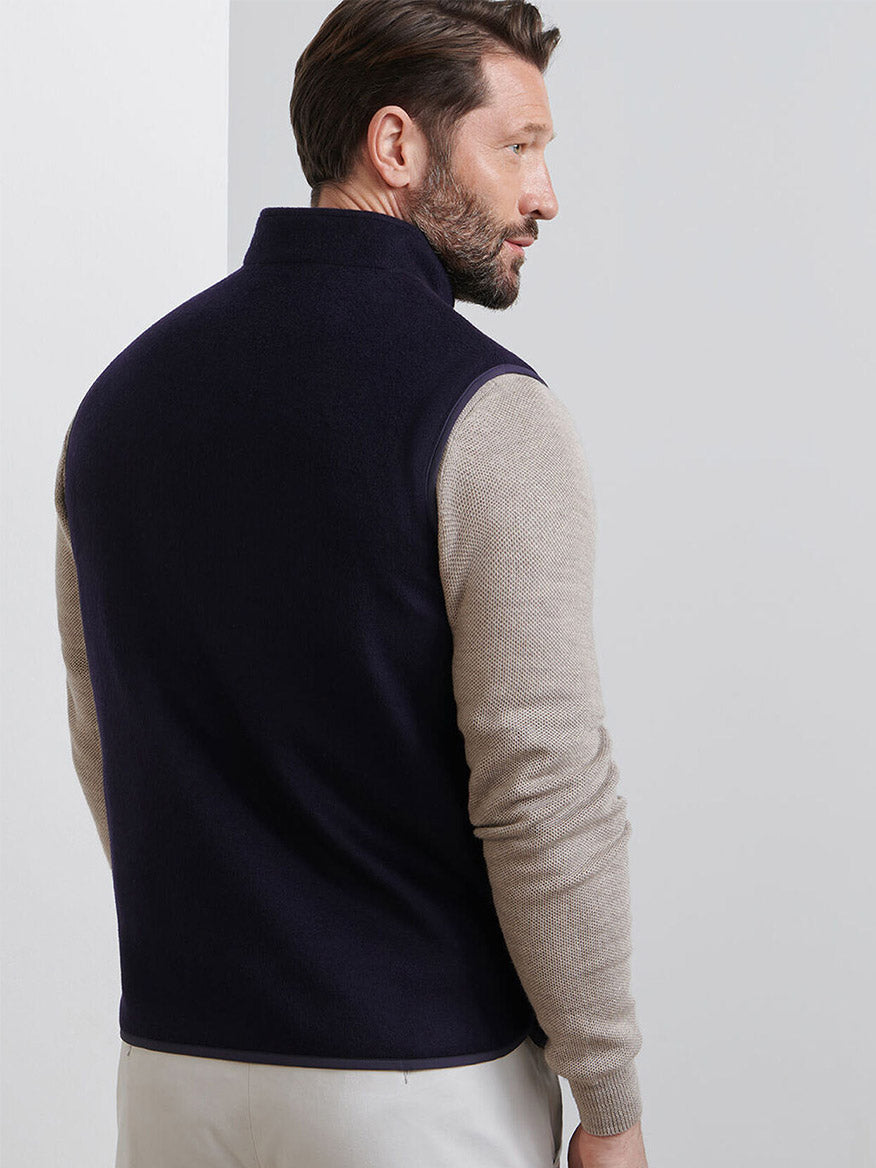 A man with brown hair and a beard wears the Peter Millar Crown Flex Merino Vest in Navy over a beige long-sleeve shirt and light pants, standing with his back turned.