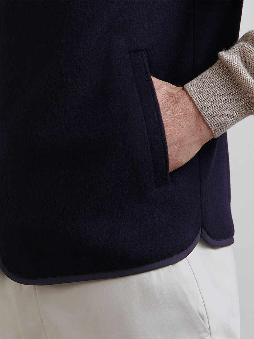 A person’s hand is placed inside the pocket of a navy Peter Millar Crown Flex Merino Vest, styled with a beige long-sleeve shirt and light-colored pants.