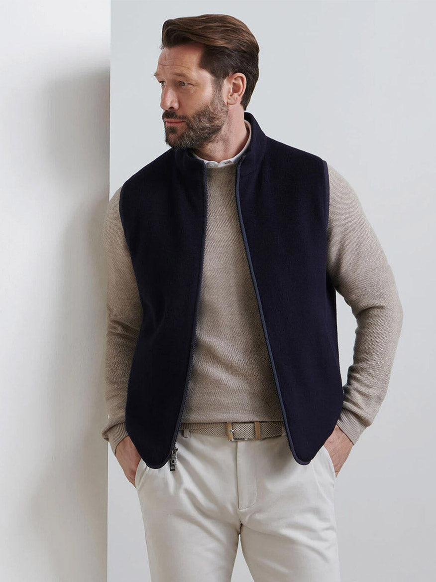 A man stands against a white wall, wearing a beige sweater, cream pants, and the Peter Millar Crown Flex Merino Vest in Navy—an effortlessly versatile layer—with one hand in his pocket, looking to his left.