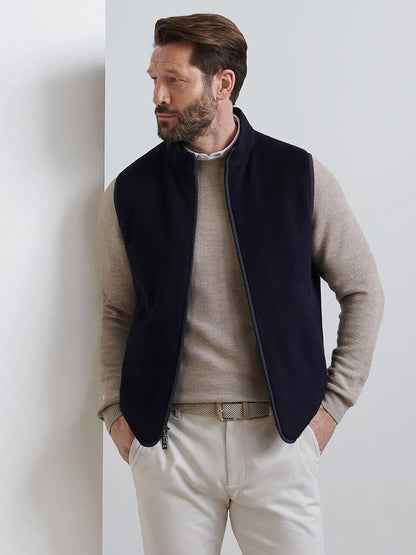 A man stands against a white wall, wearing a beige sweater, cream pants, and the Peter Millar Crown Flex Merino Vest in Navy—an effortlessly versatile layer—with one hand in his pocket, looking to his left.