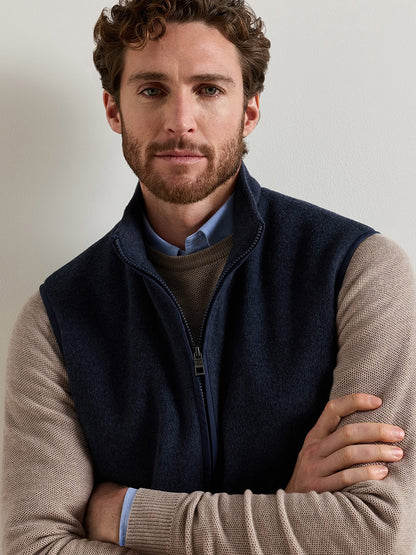 A man wearing a Peter Millar Crown Flex Merino Vest in Atlantic Blue over a light brown sweater and a light blue collared shirt.