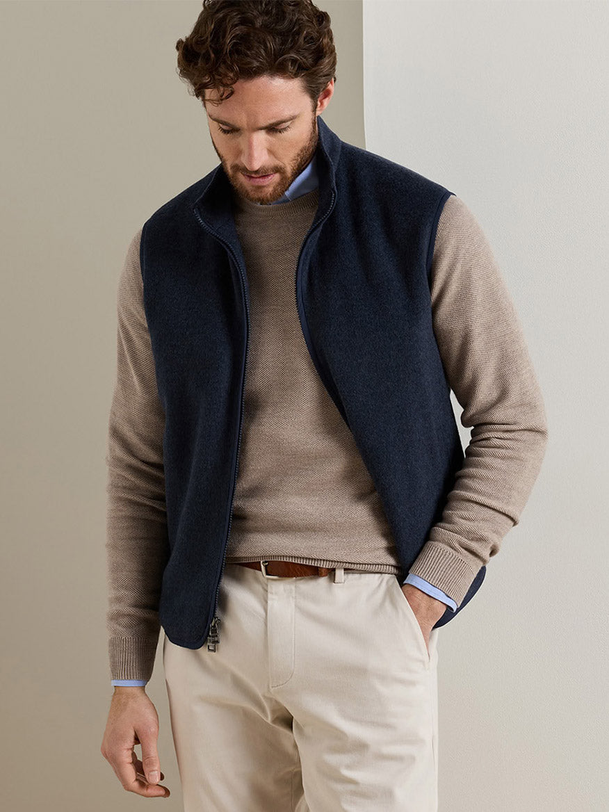 A man wearing a Peter Millar Crown Flex Merino Vest in Atlantic Blue over a tan crew-neck sweater, light blue button-down shirt, and beige trousers.