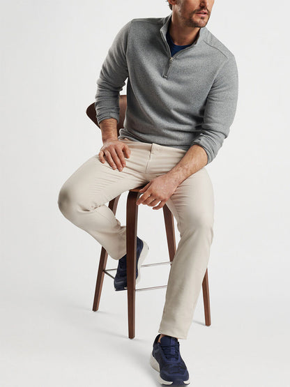 A man sits on a wooden stool, wearing the Peter Millar Crown Sweater Fleece Quarter-Zip in Gale Grey paired with beige pants and blue sneakers, set against a plain background.