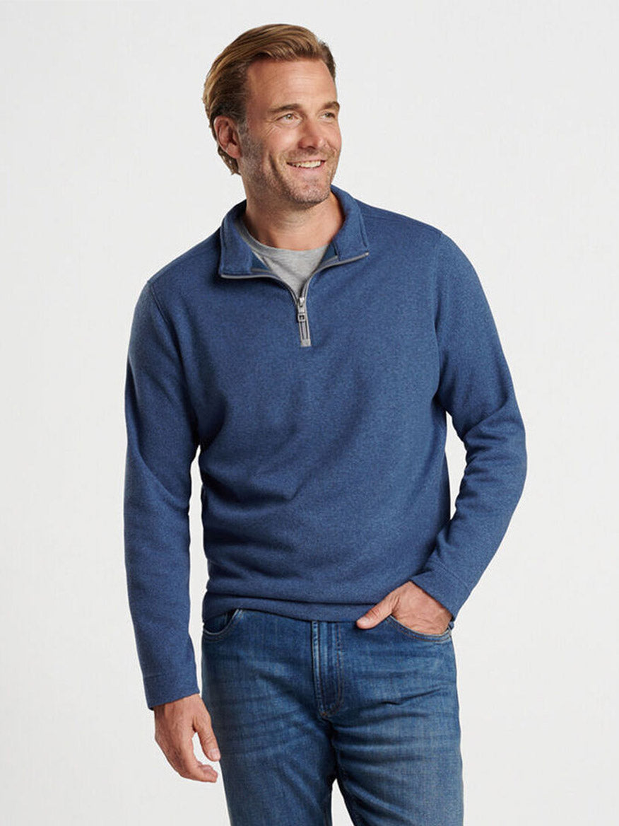 A man wearing the Peter Millar Crown Sweater Fleece Quarter-Zip in Ocean Blue, made from recycled materials, and jeans stands against a plain background, looking to the side and smiling.