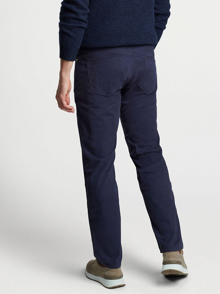 A person wearing the Peter Millar Delvin Twill Five-Pocket Pant in navy, paired with a dark, perfectly tailored sweater, stands on a light gray background, shown from the back.