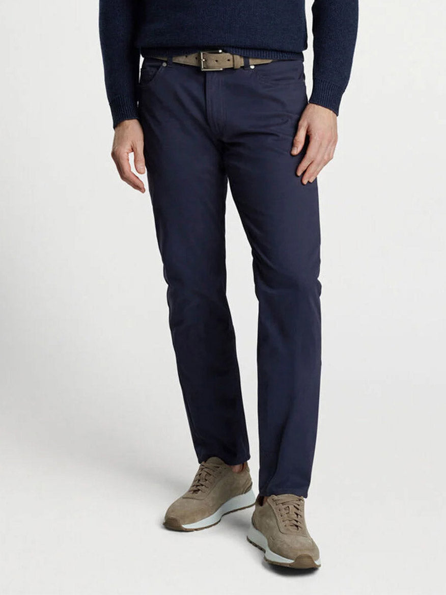 A person in Peter Millar Delvin Twill Five-Pocket Pants in navy, paired with a navy sweater and light brown shoes, stands against a plain background.