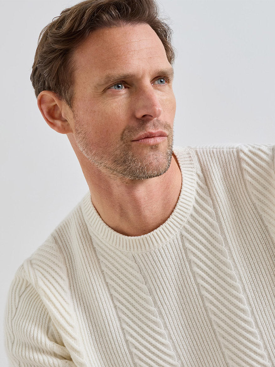 A man wearing a Peter Millar Dolomite Crewneck Sweater in Almond, crafted from cream-colored merino-cashmere with a textured diagonal ribbed pattern.