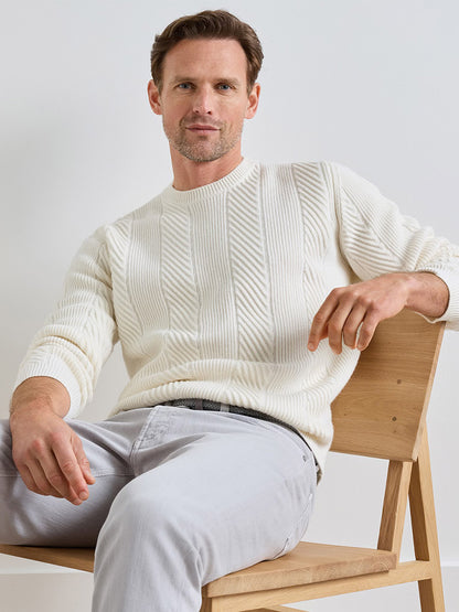 A man wearing a cream-colored, textured diagonal rib Peter Millar Dolomite Crewneck Sweater in Almond, seated on a wooden chair.