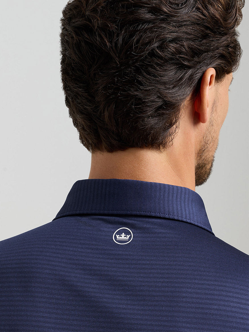 A man is seen from behind wearing the Peter Millar Empire Performance Jersey Polo in navy, featuring a small white crown logo below the collar and built-in UPF 50+ sun protection.