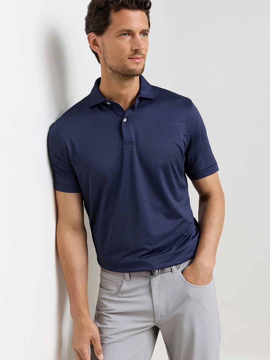 A man wearing the Peter Millar Empire Performance Jersey Polo in Navy and light gray pants stands against a white wall, one hand in his pocket.