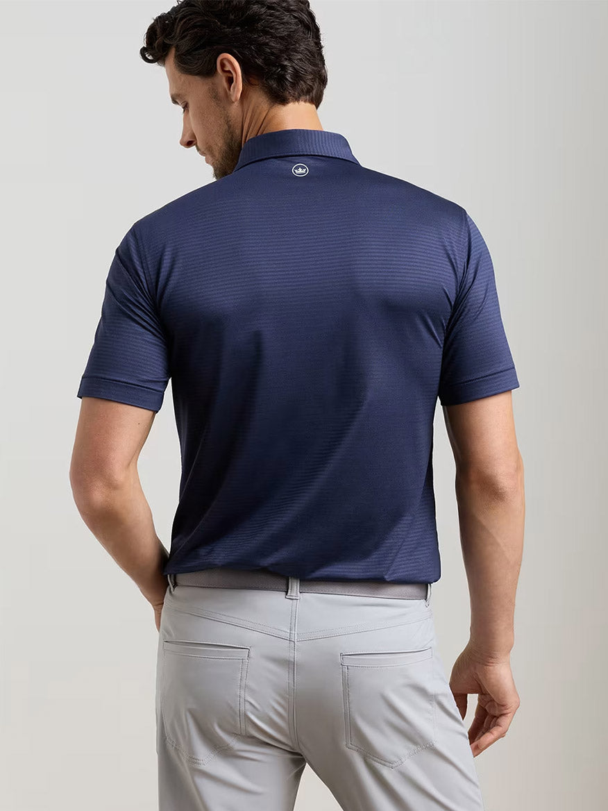 A man faces away, wearing the Peter Millar Empire Performance Jersey Polo in Navy (UPF 50+) and light gray pants against a plain light background.