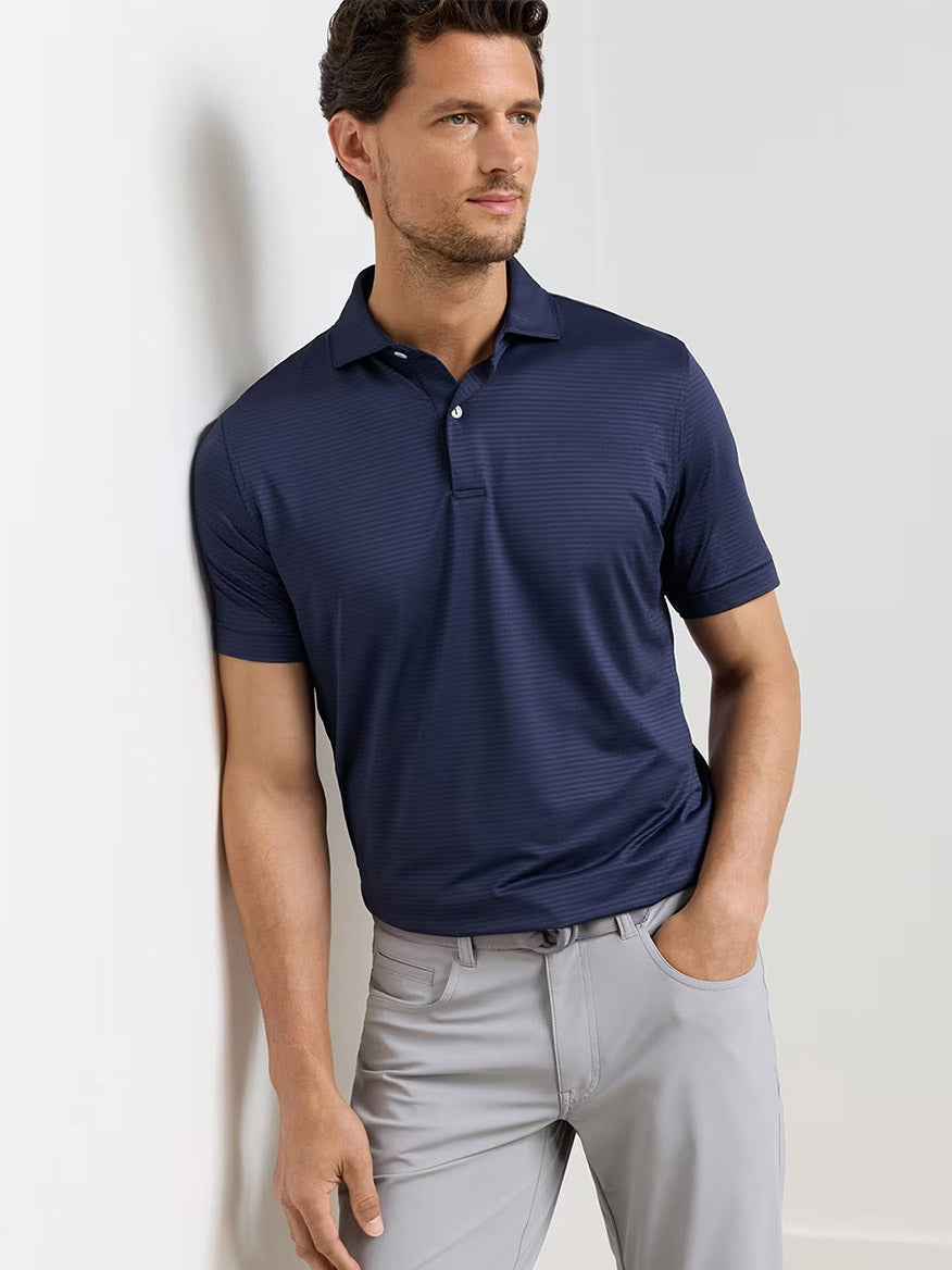 A man wears the Peter Millar Empire Performance Jersey Polo in Navy and light gray pants, standing against a plain, light background with one hand in his pocket.