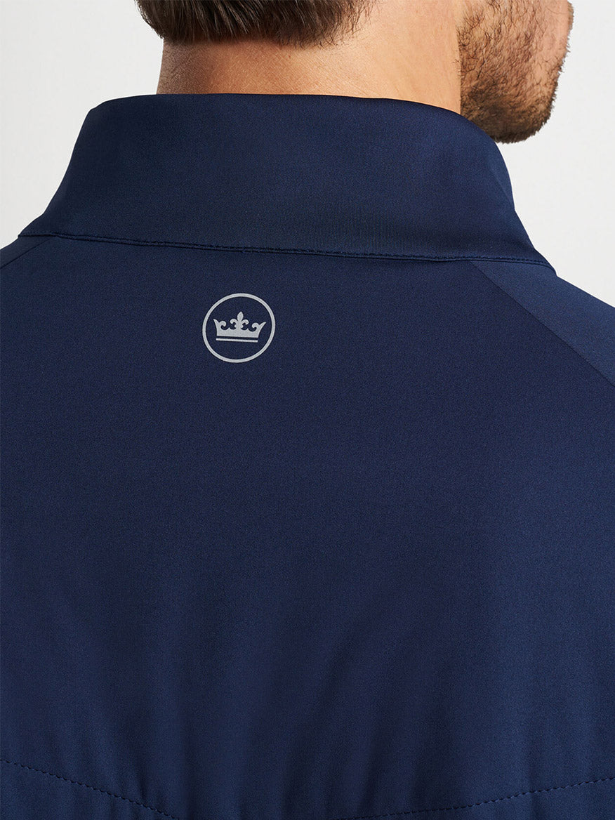 A close-up of a man's nape wearing a Peter Millar Merge Hybrid Jacket in Navy, crafted from performance fabric, offering lightweight warmth and water-resistant capabilities, with a small logo featuring a crown on the back of the collar.