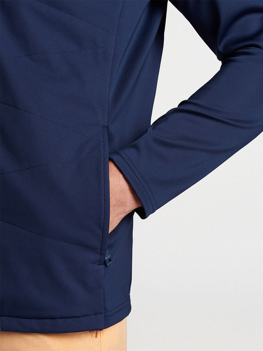 A close-up of a person wearing a Peter Millar Merge Hybrid Jacket in Navy, with their hand placed on their hip, showcasing the jacket's seam and lightweight warmth detail.