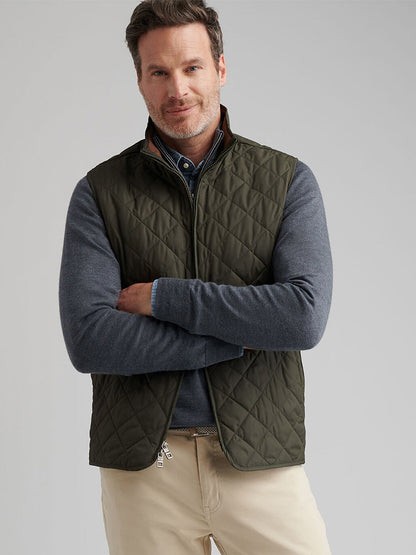 A man with short hair and a trimmed beard stands with arms crossed against a plain background, wearing the Peter Millar Essex Vest in Dark Olive over a gray long-sleeve shirt and beige pants.