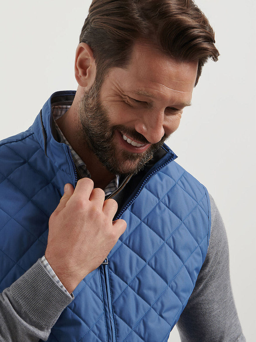 A man with brown hair and a beard smiles while zipping up a Peter Millar Essex Vest in Astral Blue over a grey long-sleeve shirt.