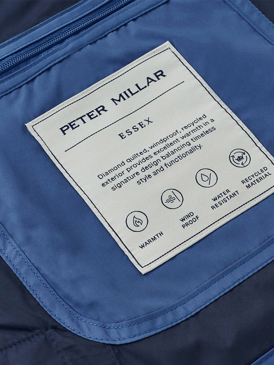 Close-up of the Peter Millar Essex Vest in Astral Blue label, highlighting its warmth, windproof and water-resistant features, and construction from recycled materials on blue quilted fabric.