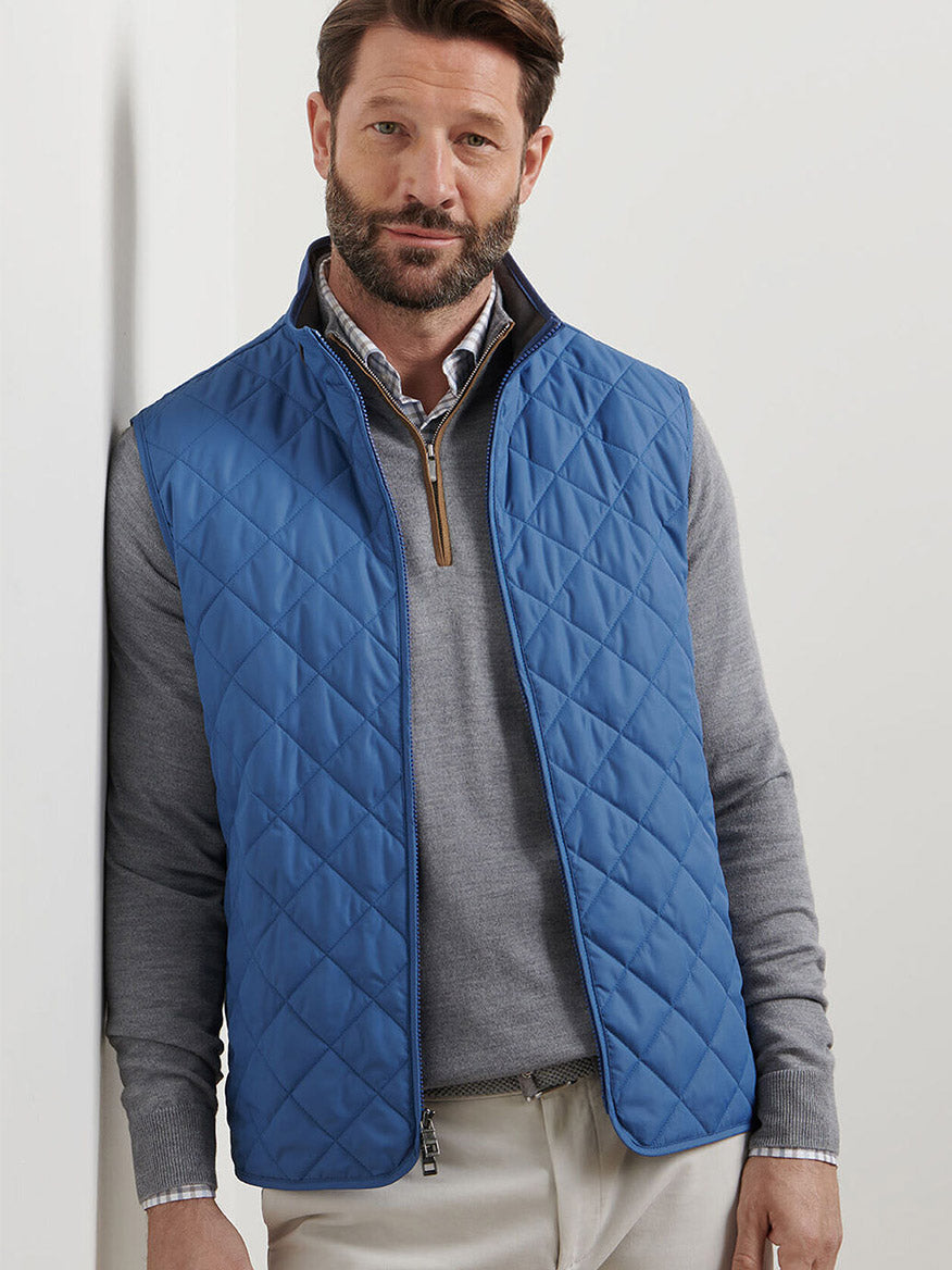 Man wearing the Peter Millar Essex Vest in Astral Blue over a light gray sweater and collared shirt, standing against a white wall and looking at the camera.