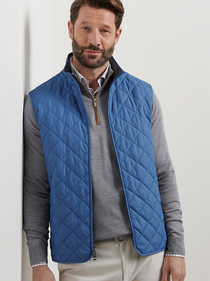 Man wearing the Peter Millar Essex Vest in Astral Blue over a light gray sweater and collared shirt, standing against a white wall and looking at the camera.
