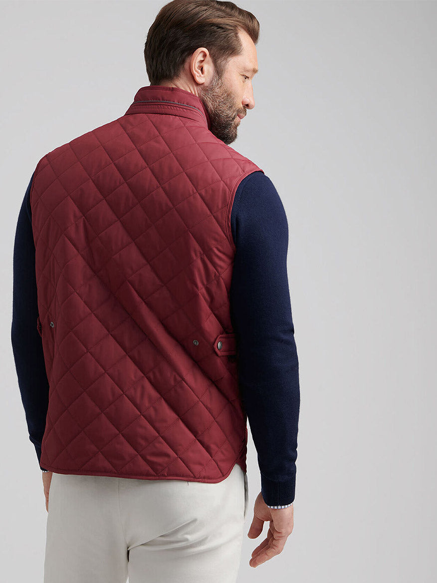 A man wears the Peter Millar Essex Vest in Autumn Foliage over a navy sweater and white pants, standing with his back to show off the water-resistant, quilted vest.
