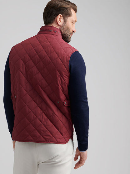 A man wears the Peter Millar Essex Vest in Autumn Foliage over a navy sweater and white pants, standing with his back to show off the water-resistant, quilted vest.