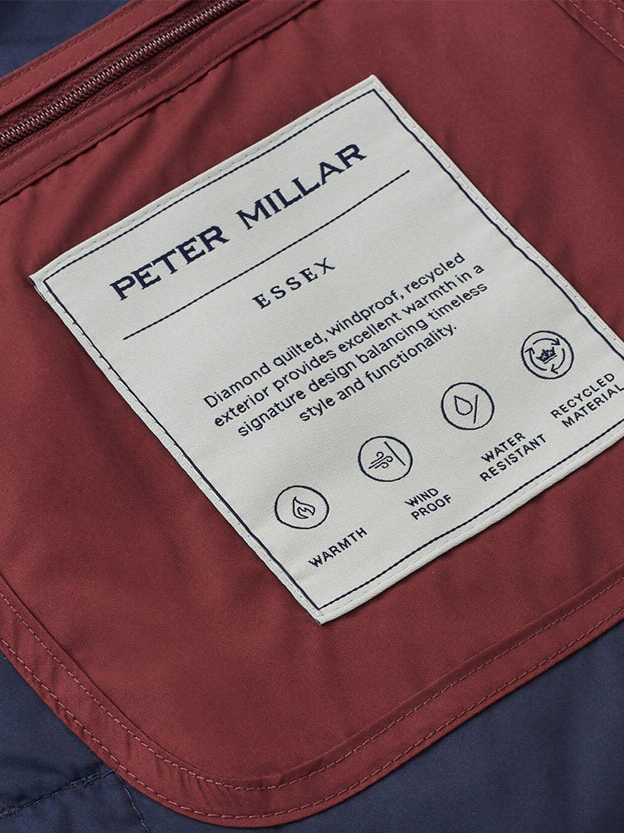 A close-up of the Peter Millar Essex Vest in Autumn Foliage label, highlighting its windproof, waterproof, and recycled material features, sewn onto maroon and navy fabric.